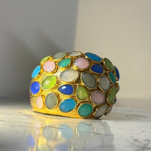Könnte beinhalten: Ein goldfarbenes Armband mit einer Vielzahl farbiger, oval geformter Edelsteine. Die Edelsteine sind in den Farbtönen Rosa, Blau, Grün und Weiß gehalten und bilden ein lebendiges Design. Das Armband hat eine strukturierte Oberfläche. Das Armband ist ca. 5 cm breit.