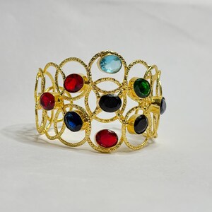 Pode incluir: Pulseira de punho dourada com um design de círculos interligados. É adornada com pedras preciosas ovais em vermelho, azul, verde e preto. As pedras são engastadas na estrutura dourada.