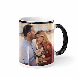 Può includere: Tazza in ceramica bianca con manico e bordo neri. La tazza presenta una foto a colori di una coppia, con l'uomo che bacia la fronte della donna. La donna tiene in mano un mazzo di fiori rosa e pesca.
