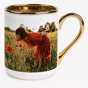 Può includere: Una tazza in ceramica bianca con una stampa fotografica di una persona con un vestito rosso a pois in un campo di papaveri. La tazza presenta un bordo e un manico in metallo dorato, che conferiscono una finitura riflettente al design.