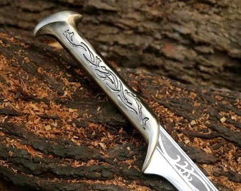 May include: A metallic knife with a polished silver-toned handle featuring intricate, dark, flowing engraved patterns. The blade also displays matching decorative etched designs along its surface, extending from the hilt toward the point.