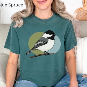 May include: A blue spruce colored t-shirt featuring a graphic illustration of a black-capped chickadee perched on a branch. The bird is positioned in front of two overlapping circles in muted olive green and light blue. The text Blue Spruce is visible.