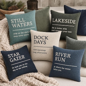 May include: A collection of square throw pillows in various colors including teal, olive green, cream, and navy blue. Each pillow features white or dark text with phrases like Still Waters, Lakeside, Dock Days, Star Gazer, and River Run.