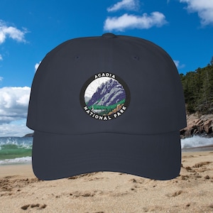 May include: A navy blue baseball cap featuring a circular embroidered patch on the front. The patch depicts a mountain landscape with green trees and purple rocky peaks, surrounded by the text Acadia National Park in white capital letters.