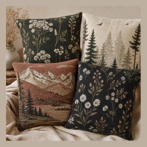 May include: Four square throw pillows featuring woven designs. Two pillows display dark charcoal fabric with white wildflower patterns. One pillow shows a cream background with green pine trees. One pillow depicts a mountain landscape in rust tones.