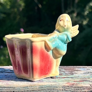 May include: A vintage ceramic planter with a rectangular shape and a red and yellow color scheme. The planter features a small angel figure with blue accents. The planter is likely used for small plants or flowers.