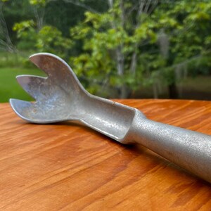 May include: A silver-colored metal ice cream scoop with a textured, matte finish. The scoop features a wide, three-pronged head designed for portioning frozen desserts, connected to a cylindrical handle that tapers toward the base.