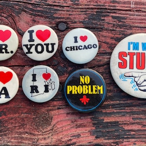 May include: A collection of vintage round buttons with various designs and text. Some buttons say "I LOVE" with a heart and the text "J.R.", "YOU", "LA", "Chicago", and "I RI". One button says "NO PROBLEM", and another says "I'M WITH STUPID".
