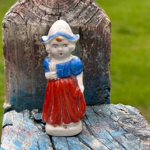 May include: A small ceramic figurine of a girl in traditional Dutch attire. The figurine wears a white bonnet with a blue trim, a blue top, and a red skirt. The figure is standing on a weathered wooden surface.