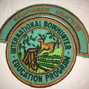 May include: Embroidered patch with teal and brown accents. The patch features the text "INTERNATIONAL BOWHUNTER EDUCATION PROGRAM" and "QUALIFIED BOWHUNTER". A deer is depicted in a forest scene.