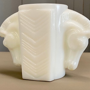 May include: A white, opaque glass vase with a geometric design. The vase features two ram's head sculptures on either side of a rectangular body with a chevron pattern. The vase has a rounded base and a wide opening.