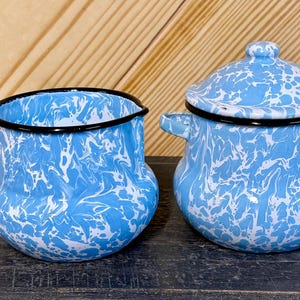 May include: A blue and white enamelware creamer and covered pot set. The creamer has a handle and black trim. The covered pot has two handles and a lid with a knob. Both pieces have a blue and white swirled pattern.