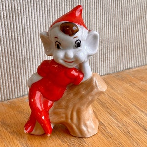 May include: A ceramic figurine of a smiling elf wearing a red outfit and hat. The elf is leaning against a tan-colored tree stump. The figurine has white skin, large ears, and brown hair peeking out from under the hat.