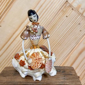 May include: A handcrafted seashell figurine of a woman. The figure has a black head, a shell torso, and a skirt made of white shells. The arms are made of white wire and shell accents. The figurine is decorated with orange and brown shells.