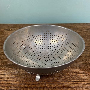 May include: A round, silver-colored metal colander featuring a wide, open top and a perforated bowl design. The kitchen tool is supported by small, integrated metal feet at the base, allowing it to sit elevated on a flat surface.