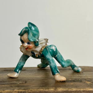 May include: A ceramic figurine of a crawling elf with a marbled green and white finish. The elf wears a pointed green hat, a white ruffled collar with gold trim, and has painted facial features including blue eyes and rosy cheeks.