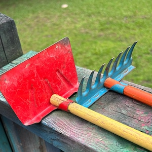 May include: A red garden hoe and a blue rake with wooden handles rest on a weathered wooden surface. The hoe has a rectangular blade, and the rake features curved tines. The tools are designed for gardening.