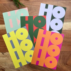Può includere: Una collezione di biglietti d'auguri colorati con la scritta "HO HO HO". I biglietti sono in vari colori, tra cui verde, giallo, arancione e viola, con le lettere in colori contrastanti. Sono disposti su una superficie di legno.