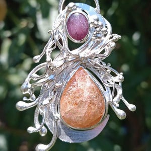 Puede incluir: Colgante de plata con una gema naranja en forma de lágrima y una gema púrpura ovalada más pequeña. El colgante tiene un diseño ornamental y fluido con detalles de hojas y vides, suspendido de un lazo plateado.