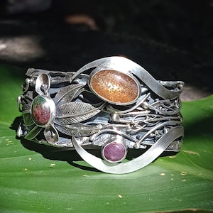 Op de afbeelding: Een brede, sierlijke zilveren armband met drie ovale edelstenen: een grote, sprankelende amberkleurige steen, een kleinere rode steen en een groen-rode steen. De armband heeft een gedetailleerd, door de natuur geïnspireerd ontwerp met bladeren en ranken.