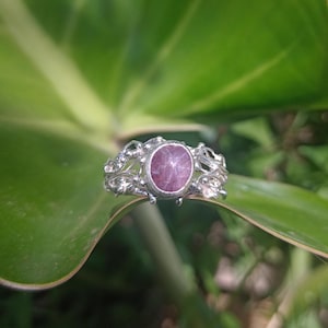 Puede incluir: Un anillo de plata con una gema ovalada de color rosa púrpura. El anillo tiene un diseño calado y ornamentado. El anillo se encuentra sobre un fondo de hojas verdes, creando un contraste natural.