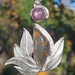 May include: A silver pendant necklace. The pendant features a large, teardrop-shaped stone with orange and brown inclusions, set within a silver leaf design. Above, a smaller, round purple stone is set in a swirling silver setting. The necklace hangs on a silver chain.