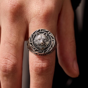 May include: A silver-toned ring featuring a detailed wolf head design at the center. The ring has an intricate border with geometric patterns. The ring is worn on a finger, showcasing its metallic sheen and craftsmanship.