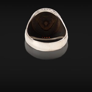 May include: A silver ring with a dark brown interior and a decorative design. The ring has a smooth, polished finish and a detailed pattern around the top edge. Four small orange stones are set in the ring.