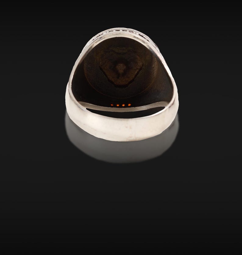 May include: A silver ring with a dark brown interior and a decorative design. The ring has a smooth, polished finish and a detailed pattern around the top edge. Four small orange stones are set in the ring.