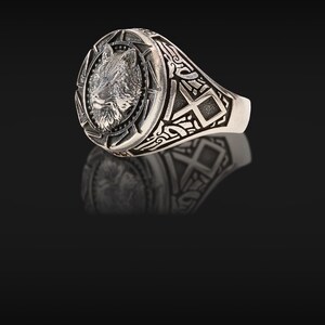 May include: A silver ring featuring a detailed wolf head design. The ring has an oval face with a textured wolf portrait surrounded by a starburst pattern. The band is adorned with intricate geometric and scrollwork patterns.
