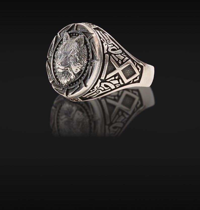 May include: A silver ring featuring a detailed wolf head design. The ring has an oval face with a textured wolf portrait surrounded by a starburst pattern. The band is adorned with intricate geometric and scrollwork patterns.