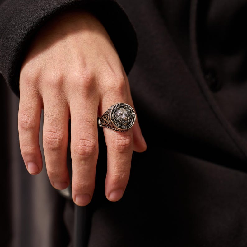 May include: A silver-toned ring featuring a detailed wolf head design at the center. The ring has an ornate, patterned border and is worn on a finger. The background is a dark, solid color.
