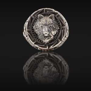 May include: A silver-colored ring featuring a detailed wolf head design at the center. The ring has an intricate border with geometric patterns and small, dark accents. The ring is displayed on a reflective black surface.