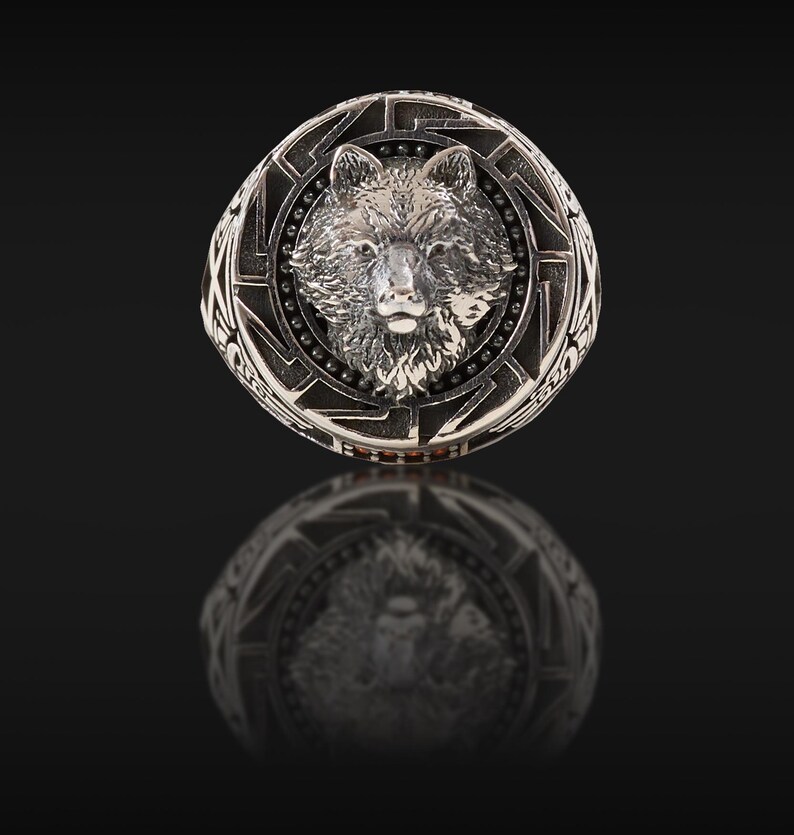 May include: A silver-colored ring featuring a detailed wolf head design at the center. The ring has an intricate border with geometric patterns and small, dark accents. The ring is displayed on a reflective black surface.