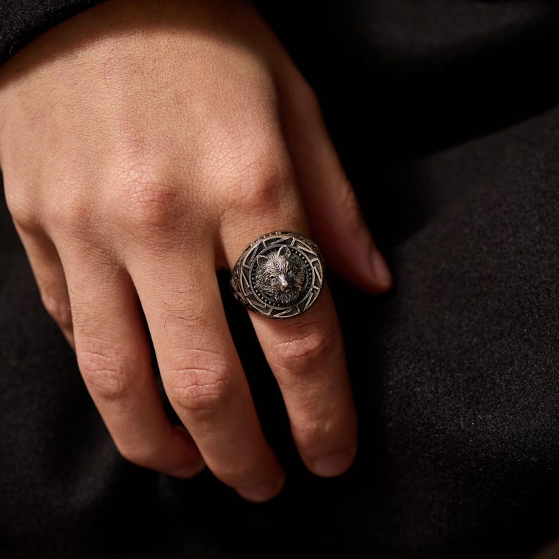 May include: A silver-colored ring featuring a detailed wolf head design. The ring has an intricate border pattern and is worn on a finger. The ring is a piece of jewelry.
