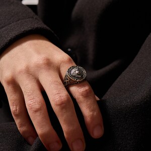 May include: A silver-toned ring featuring a detailed wolf head design at the center. The ring has an intricate border with geometric patterns. The ring is worn on a finger, showcasing its craftsmanship and style.