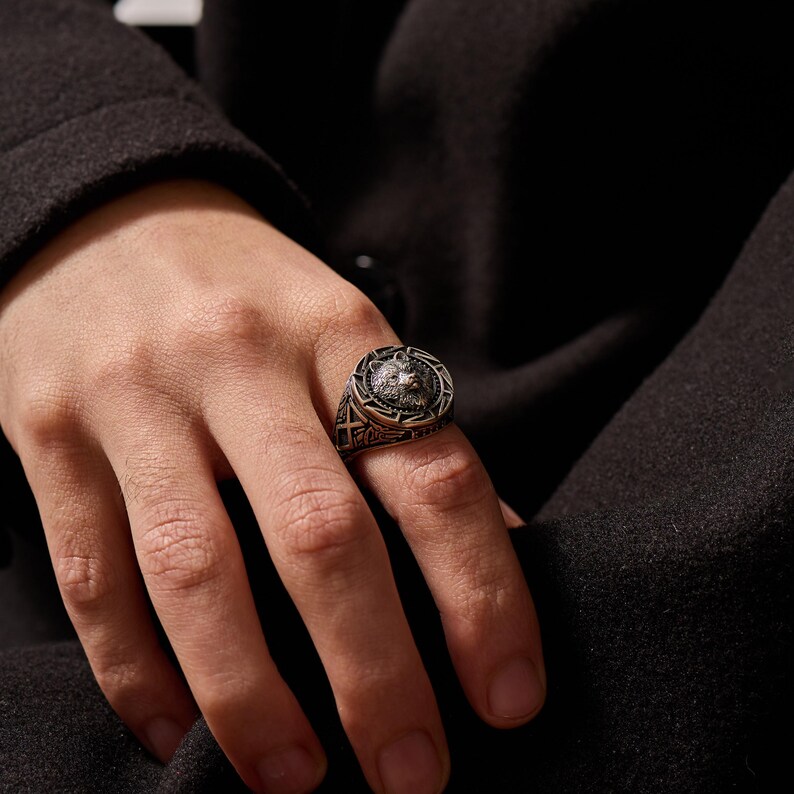 May include: A silver-toned ring featuring a detailed wolf head design at the center. The ring has an intricate border with geometric patterns. The ring is worn on a finger, showcasing its craftsmanship and style.