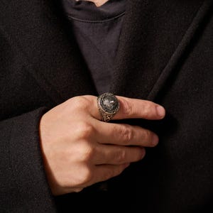 May include: A silver-toned ring with a detailed design featuring a central emblem and intricate patterns. The ring is worn on a finger, showcasing its ornate craftsmanship and metallic sheen. The ring's design includes a central emblem and detailed patterns.