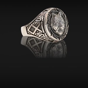 May include: Silver ring featuring a detailed wolf head design within a circular frame. The ring has an oval face and intricate patterns on the band. The ring is made of a shiny, metallic material.
