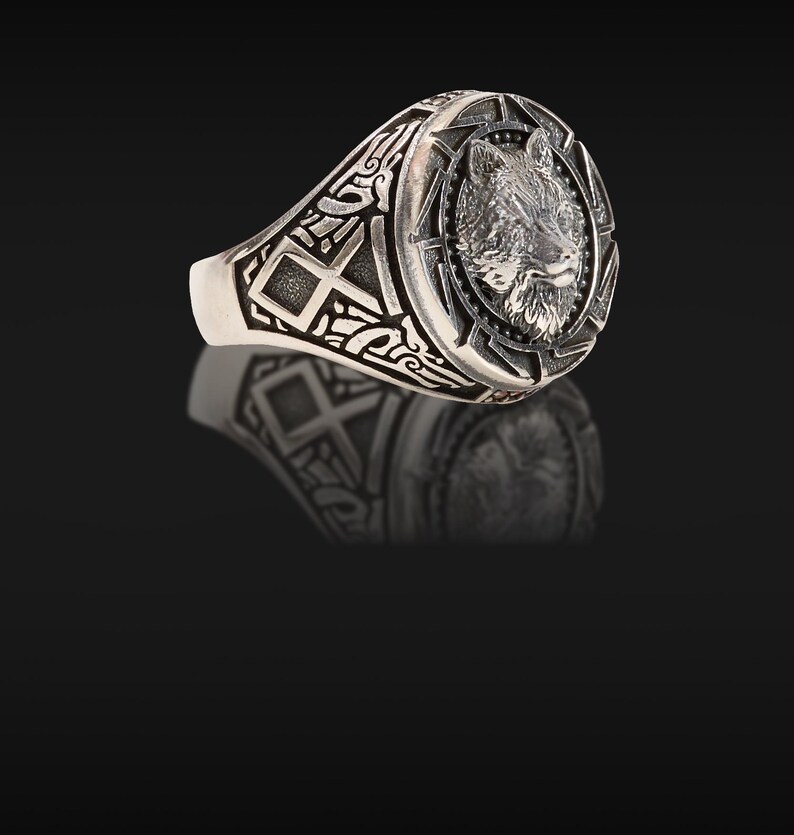 May include: Silver ring featuring a detailed wolf head design within a circular frame. The ring has an oval face and intricate patterns on the band. The ring is made of a shiny, metallic material.