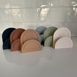 Peut inclure: Une collection de dessous de verre semi-circulaires en forme d'arche, de différentes couleurs sourdes, dont le bleu marine, le marron, le beige et le vert sauge. Chaque dessous de verre a un fin contour plus foncé. Les dessous de verre sont disposés en rangée.