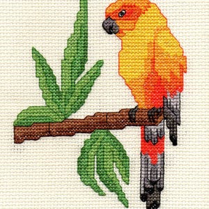May include: A cross-stitch embroidery on cream-colored fabric featuring a sun conure parrot with yellow, orange, and red feathers perched on a brown branch. The design includes green leaves and the parrot has a dark gray beak and tail feathers.