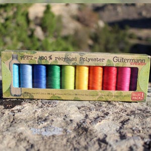 Pode incluir: Uma caixa de cartão retangular contendo dez bobines de linha de costura num gradiente de cores arco-íris. A embalagem está rotulada como rPET 100% recycled Polyester, Gütermann creativ e Value Pack, com 100 metros de linha por bobine.