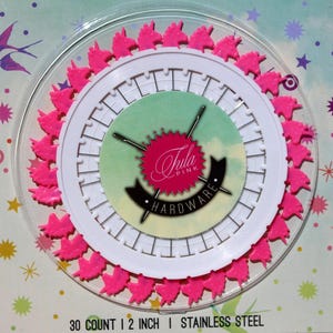May include: A circular package containing 30 count 2 inch stainless steel pins with bright pink unicorn-shaped heads. The center label features the Tula Pink Hardware logo with two crossed needles. The packaging has a light blue background with stars.