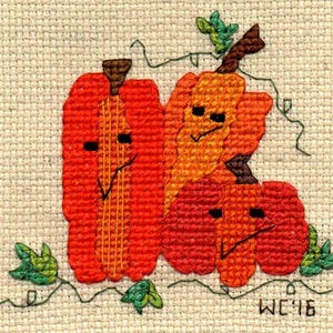 Puede incluir: Diseño de punto de cruz con tres calabazas sonrientes en tonos naranja y rojo, con vides y hojas verdes. Las calabazas tienen ojos negros y sonrisas cosidas. El diseño está sobre un fondo de tela beige. Las iniciales "WC'16" están cosidas en la esquina inferior derecha.
