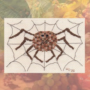 Puede incluir: Un diseño de punto de cruz con una araña marrón sobre una telaraña. La araña tiene un cuerpo texturizado y ocho patas, sobre un fondo de telaraña cosida. El diseño está sobre tela color crema, con las iniciales "WC 20" en la esquina.