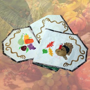May include: Three white quilted placemats with brown trim and decorative stitching. Each mat features a different autumn-themed applique: a collection of fruits and vegetables, a turkey, and fall leaves. The background is a blurred image of fall colors.