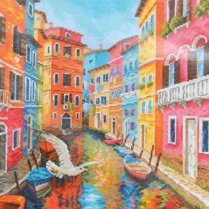 May include: A colorful cross-stitch depicting a canal scene with buildings in shades of orange, pink, blue, and yellow. Seagulls fly above the water, and small boats are docked along the canal. The water reflects the vibrant colors of the buildings.