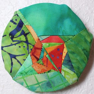 May include: A handmade fabric coaster with a patchwork design. The coaster is a mix of green, orange, red, and blue fabrics. The geometric design is made up of various shapes and patterns, with visible stitching.