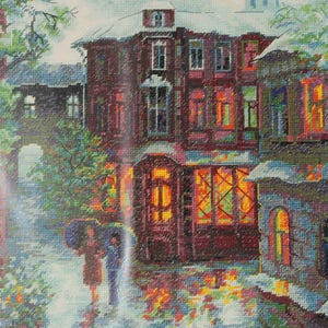 May include: A cross-stitch embroidery depicting a multi-story building with dark brown walls and illuminated windows. Two figures holding umbrellas walk along a street in the foreground. The scene includes green trees, a stone archway, and reflections on the ground.